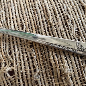 May include: Close-up of a silver fork with a decorative floral design on the handle. The silverware is set against a textured, woven background, highlighting the intricate details of the utensil.