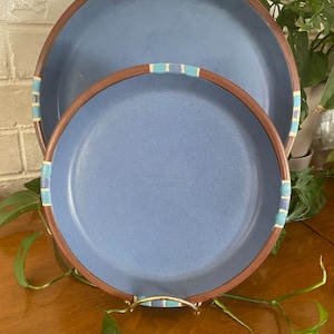 May include: Two blue ceramic plates, one large and one smaller, with brown rims and decorative blue and white accents. The plates are displayed on a wooden surface, with greenery in the background.