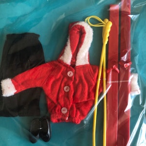 Puede incluir: Un conjunto de esquí de muñeca vintage, con una chaqueta roja con ribete blanco, pantalones negros y zapatos negros. El conjunto incluye esquís rojos y bastones de esquí amarillos. La chaqueta tiene botones blancos y una capucha con ribete blanco.