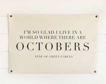 May include: A beige canvas banner with black text that reads "I'm so glad I live in a world where there are Octobers. Anne of Green Gables." The banner is hanging on a white wood wall.