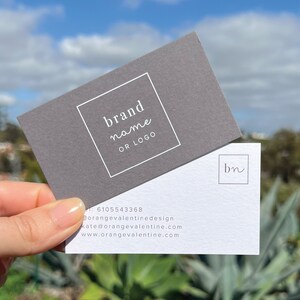 May include: Two business cards with a gray and white color scheme. The top card has a white square with the text "brand name or logo". The bottom card has a white square with the text "bn" in cursive.