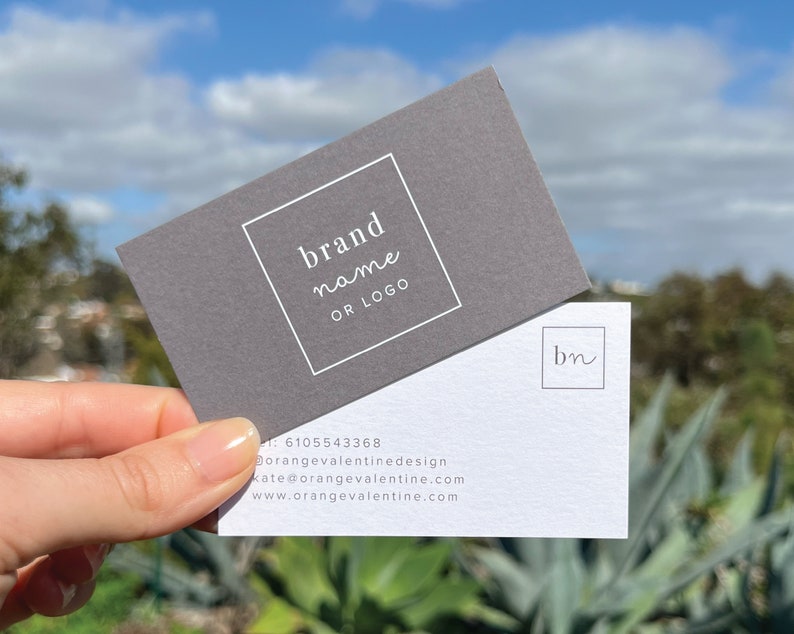 May include: Two business cards with a gray and white color scheme. The top card has a white square with the text "brand name or logo". The bottom card has a white square with the text "bn" in cursive.