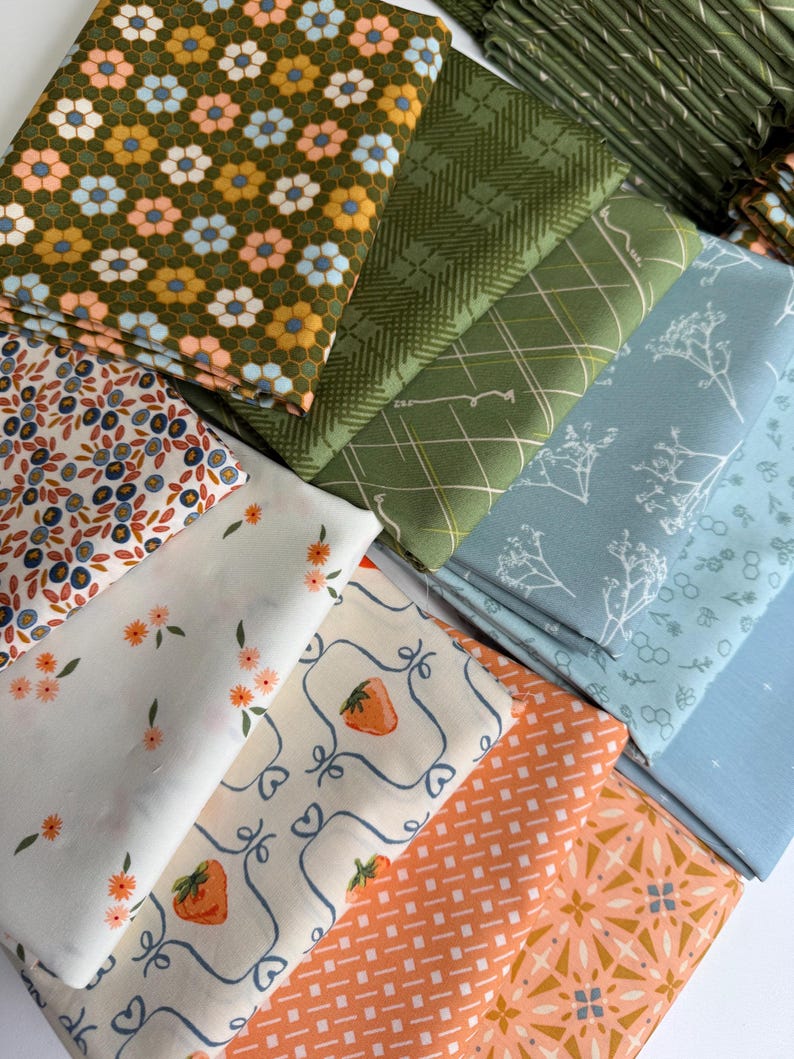 May include: A collection of folded fabric squares featuring diverse patterns including a mustard yellow honeycomb floral print, green plaid, blue botanical sketches, white with orange flowers, cream with strawberries, and orange geometric designs.