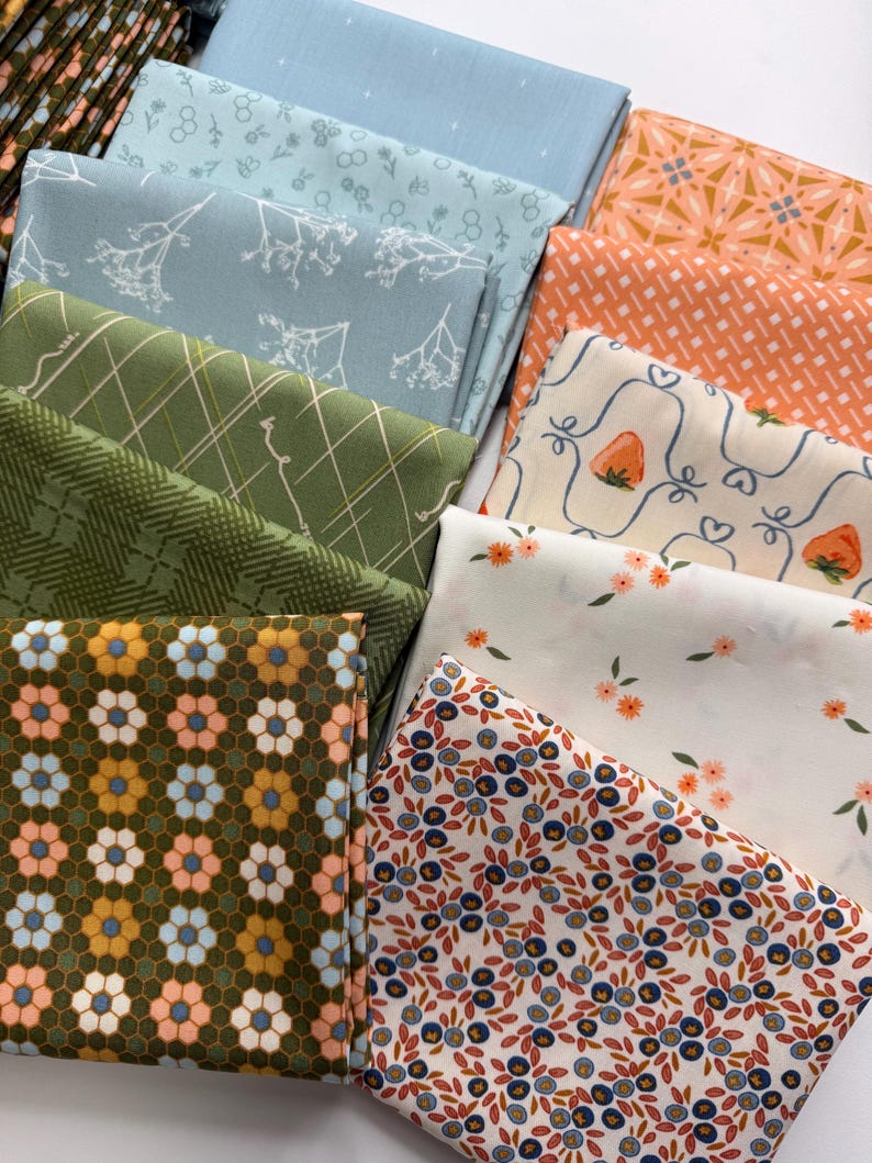 May include: A collection of folded fabric squares featuring diverse patterns including blue floral prints, green plaid, orange geometric designs, white strawberry motifs, small orange flowers, and a dark green background with repeating flower shapes.