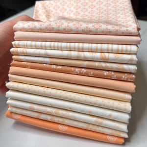 May include: A stack of fabric squares in various shades of peach, cream, and white. The fabrics feature floral, geometric, and striped patterns. The fabrics are neatly folded and stacked, showcasing a variety of designs and textures.