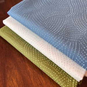 May include: A stack of three fabric squares with a white dotted pattern on a blue, beige, and green background. The fabric is folded and stacked on a wooden surface.