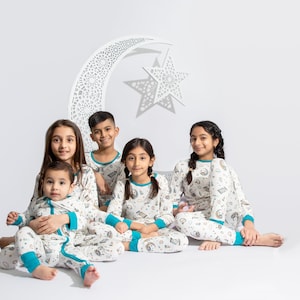 May include: Five children wearing white pajamas with a blue trim and a colorful print of stars, moons, and other celestial objects. The children are sitting in front of a white cutout of a crescent moon and star.