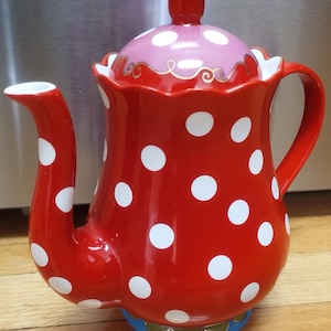Op de afbeelding: Een rode keramische theepot met witte stippen. Het deksel is roze met witte stippen en gouden rand. De theepot heeft een blauwe basis met gouden accenten. De handgreep en tuit zijn rood.