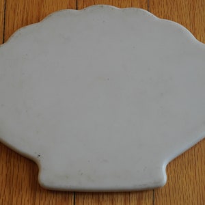 May include: A light beige, scallop-edged ceramic serving platter. The platter has a shell-like shape with a wide, flat surface and a slightly raised edge. The background is a wooden floor.