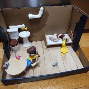 Op de afbeelding: Een miniatuur diorama in een kartonnen doos, met een kamer met een bed, toilet, wastafel en een tafel met een zittende figuur. De scène bevat kleifiguren en accessoires, met een humoristische, cartoonachtige stijl.