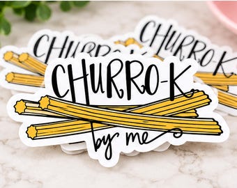 May include: A die-cut sticker featuring a graphic of three golden-brown churros arranged in a stack. The text CHURRO-K is printed in black script above the churros, and the words by me are written in black script below the churros.