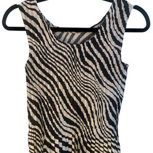 May include: A sleeveless top featuring a black and white wavy, striped pattern throughout. The garment has a round neckline and a pleated texture that creates a dynamic, rippled effect across the fabric, ending in a straight hemline.