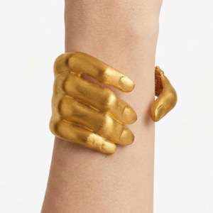 May include: A gold-toned cuff bracelet shaped like a hand wrapping around the wrist. The design features four fingers resting across the front of the wrist, with a separate thumb piece extending from the hinge on the opposite side of the band.
