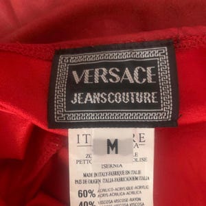 May include: A red garment featuring a black Versace Jeans Couture label with a white Greek key border. Below, a white tag displays the size M, origin as Italy, and a fabric composition of 60 percent acrylic and 40 percent viscose rayon.