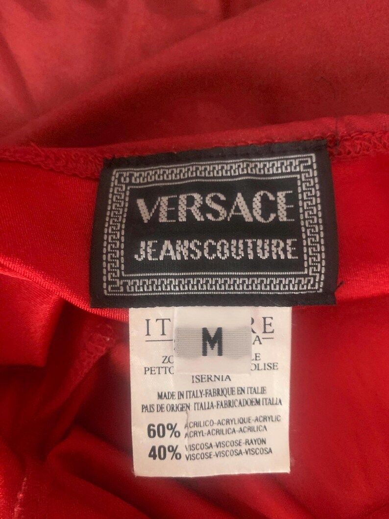 May include: A red garment featuring a black Versace Jeans Couture label with a white Greek key border. Below, a white tag displays the size M, origin as Italy, and a fabric composition of 60 percent acrylic and 40 percent viscose rayon.