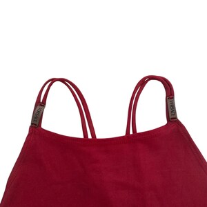 May include: A deep red top featuring a high neckline and thin double straps. Each strap is accented with a small, rectangular metallic hardware piece engraved with the word VERSACE, positioned where the straps connect to the garment body.