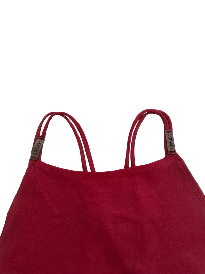 May include: A deep red top featuring a high neckline and thin double straps. Each strap is accented with a small, rectangular metallic hardware piece engraved with the word VERSACE, positioned where the straps connect to the garment body.