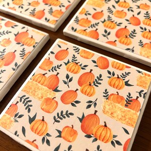 May include: Set of four ceramic coasters with a fall theme. The coasters feature a beige background with a repeating pattern of orange pumpkins, green leaves, and brown hay bales.