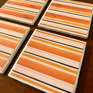 May include: Four square coasters with a watercolor-style striped design in shades of orange, peach, and black. The coasters have a white border and are arranged on a dark wooden surface. The striped pattern is horizontal.