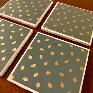May include: Set of four square coasters with a white border. The coasters are made of a dark green material with a pattern of gold acorns.