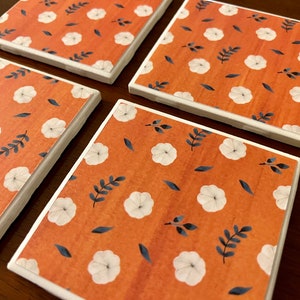 May include: Set of four square ceramic coasters with a white floral pattern on an orange background. The coasters have a white border.