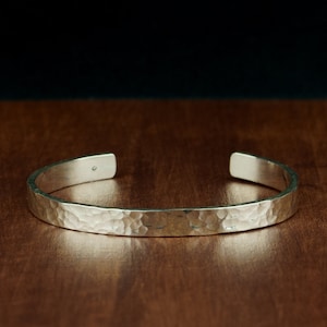 Könnte beinhalten: Ein silbernes, gehämmertes Armreif-Armband. Das Armband ist glatt und glänzend, mit einer gehämmerten Textur.