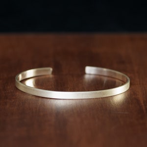 Peut inclure: Un bracelet manchette argenté avec une finition brossée. Le bracelet ouvert est présenté sur une surface en bois brun foncé, avec un fond noir. Le bracelet mesure environ 0,6 cm de large.