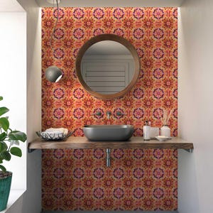 May include: A bathroom featuring a patterned wall in orange, pink, and yellow. A round wooden mirror is above a dark grey sink. A wooden shelf holds a basket, toiletries, and decorative items. A window and a hanging light fixture are also visible.