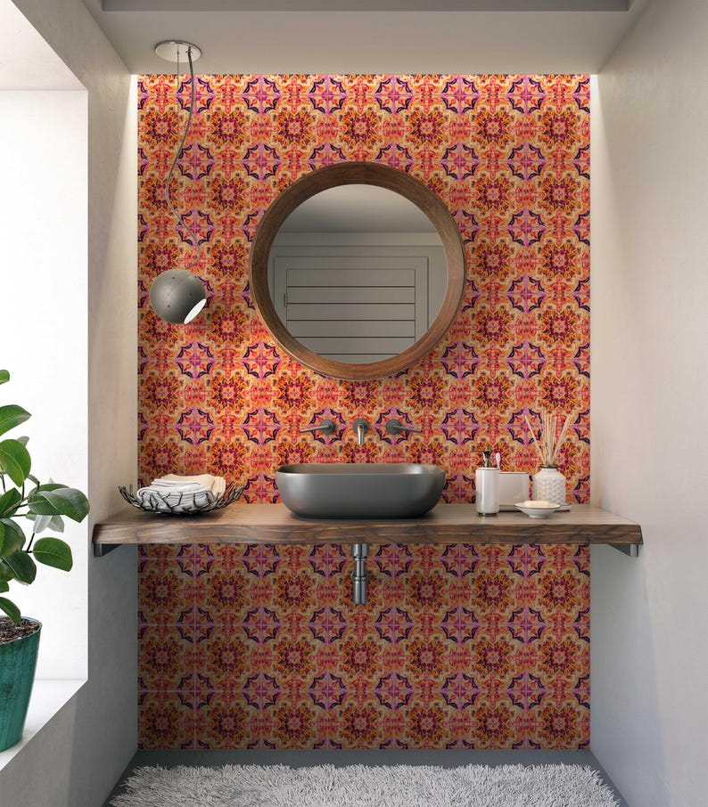 May include: A bathroom featuring a patterned wall in orange, pink, and yellow. A round wooden mirror is above a dark grey sink. A wooden shelf holds a basket, toiletries, and decorative items. A window and a hanging light fixture are also visible.