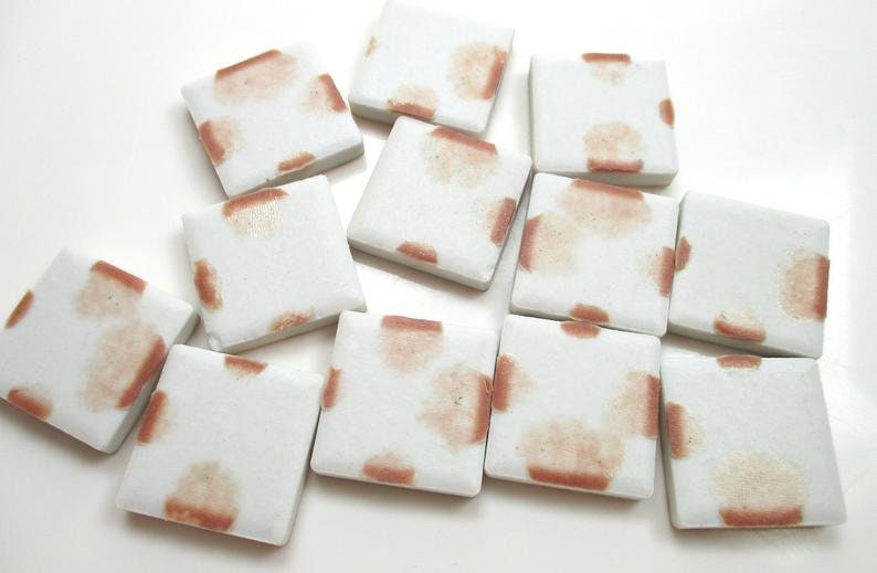 May include: A collection of white, square ceramic tiles with a reddish-brown pattern. The tiles are arranged on a white surface, showcasing their design and texture. The reddish-brown pattern appears to be a stain or a decorative element.