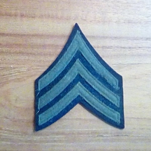 May include: A dark green military rank insignia with three stripes. The insignia is shaped like a chevron and is made of fabric.