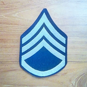 May include: A blue and white military rank patch with three silver chevrons. The patch is shaped like a teardrop.