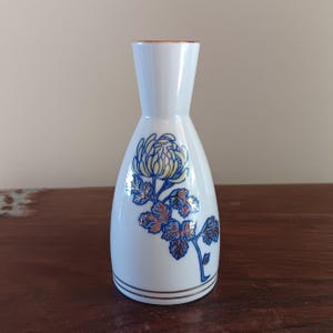 May include: White ceramic sake bottle with a floral design. The bottle features a blue and yellow chrysanthemum with orange and blue leaves. The rim and base are trimmed with gold. The bottle has a narrow neck and a rounded body.