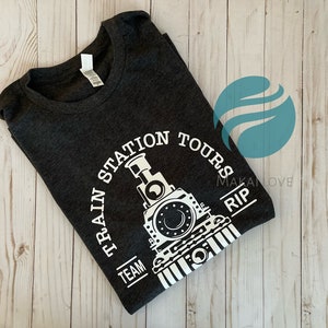 May include: A dark gray t-shirt with a white graphic of a train engine and the text "Train Station Tours Makalove RIP Team".