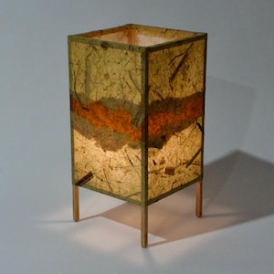 May include: A rectangular table lamp with a warm, inviting glow. The lamp features a textured, paper-like shade in shades of gold and brown, with a band of orange. It stands on four slender wooden legs, creating a soft, ambient light.