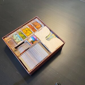 May include: A wooden game organiser with several compartments filled with game pieces, cards, and wooden dividers. The pieces are various colours, including blue, green, yellow, orange, and pink. The organiser is brown and sits on a dark surface.