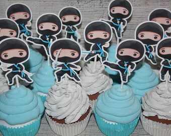 May include: Cupcakes with blue and white frosting, topped with ninja-themed paper cutouts. The ninja figures are in various action poses, wearing black outfits with blue accents and holding swords. The cupcakes are in white paper liners.