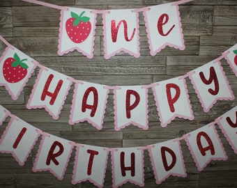 May include: A festive banner with the words "ONE HAPPY BIRTHDAY" in red glitter letters. The banner features strawberry illustrations and a scalloped pink border, strung on a pink ribbon. Perfect for a first birthday celebration.