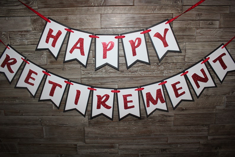 May include: A banner with the words "HAPPY RETIREMENT" in red glitter letters on white pennants with black borders. The pennants are strung on a red ribbon against a wooden background.