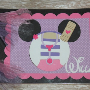 May include: A handmade scrapbook with a doctor theme. The cover features a Minnie Mouse-inspired design with a stethoscope, bandage, and the name "Willa." The design is set against a pink and purple polka dot background, with a scalloped pink border. The book is bound with a black spiral and pink tulle.