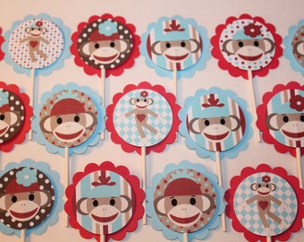 May include: A collection of cupcake toppers featuring sock monkey designs. Each topper has a scalloped red border, a light blue backing, and a wooden stick. The monkey faces are in various patterns, including stripes and polka dots, with red and blue accents.