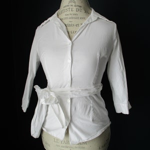 May include: A white button-down shirt with three-quarter sleeves and a tie-waist detail. The shirt is displayed on a mannequin with a black background. The shirt has a classic collar and a fitted silhouette.