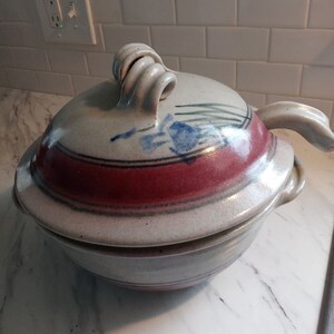 May include: A white ceramic casserole dish with a lid. The dish has a red and blue design with a swirl handle.
