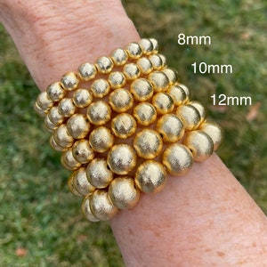 May include: A set of three gold-toned bracelets with brushed metal beads. The bracelets are stacked on a person's wrist, with the smallest bracelet measuring 8mm, the middle bracelet measuring 10mm, and the largest bracelet measuring 12mm.