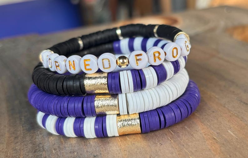 May include: A stack of four bracelets with purple, white, and black beads. The top bracelet has white beads with the word "FROG" spelled out in gold letters. The second bracelet has black beads with gold accents. The third and fourth bracelets have alternating purple and white beads.