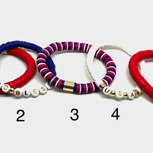 May include: Five bracelets with red, white and blue beads. The bracelets have gold accents and white beads with the words "GOD BLESS" and "USA" spelled out.