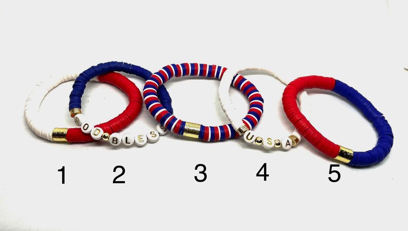May include: Five bracelets with red, white and blue beads. The bracelets have gold accents and white beads with the words "GOD BLESS" and "USA" spelled out.