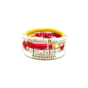 May include: Four stacked bracelets in red, white, and gold. The top bracelet is yellow, the second is red, the third is red and gold, and the bottom is white. The bracelets spell out "KANSAS CITY CHIEFS" with gold lettered beads.