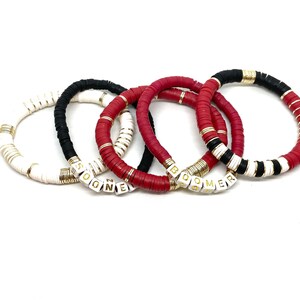 May include: Four bracelets with white and gold beads, two red bracelets with gold beads, and one black and white bracelet with gold beads. The bracelets spell out the words "Boomer" and "SOGNE".