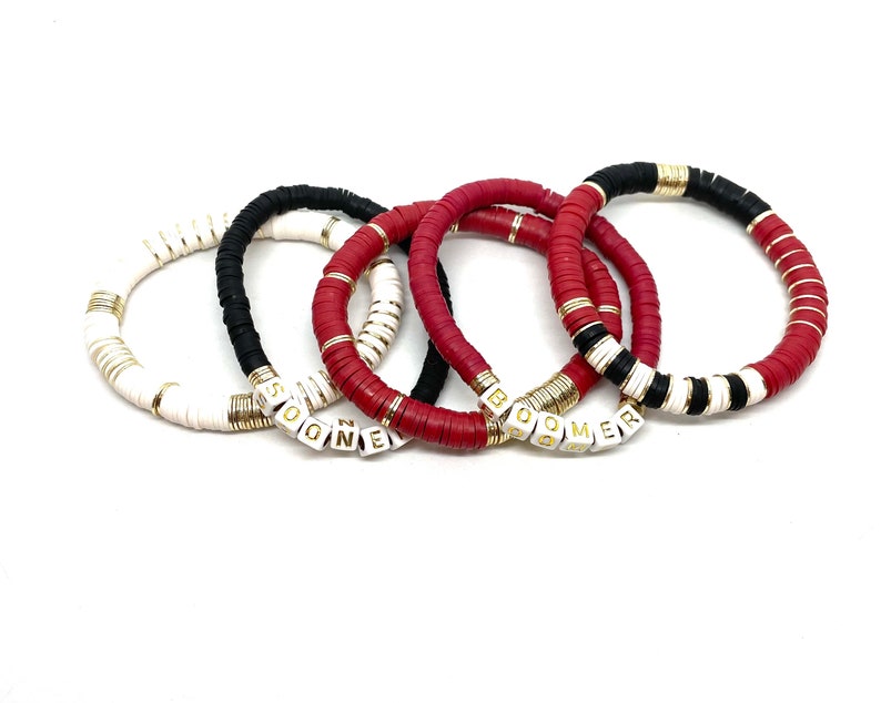 May include: Four bracelets with white and gold beads, two red bracelets with gold beads, and one black and white bracelet with gold beads. The bracelets spell out the words "Boomer" and "SOGNE".